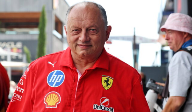 Smiling man in a red Ferrari team polo with sponsor logos (HP, Shell, UniCredit) outdoors in a paddock area.
