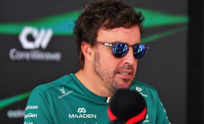 Man in teal racing jersey wearing mirrored sunglasses speaks into a microphone during an interview, race team backdrop behind him