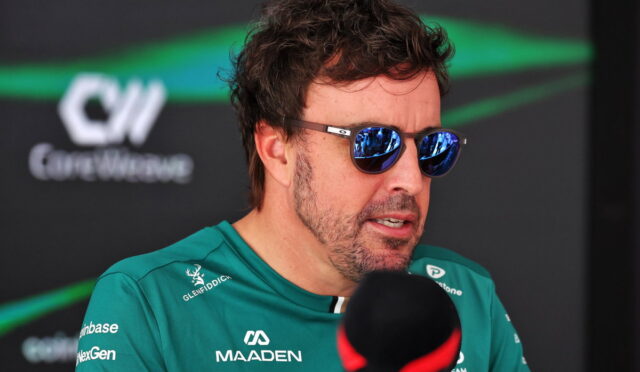 Man in teal racing jersey wearing mirrored sunglasses speaks into a microphone during an interview, race team backdrop behind him
