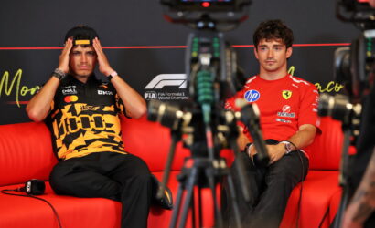 Two Formula 1 drivers sit on a red couch for an interview, cameras in the foreground.