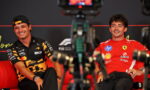 Two Formula 1 drivers sit on a red sofa during an interview, smiling, with camera gear in the foreground.