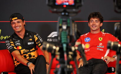Two Formula 1 drivers sit on a red sofa during an interview, smiling, with camera gear in the foreground.
