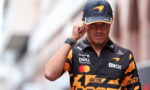 Male race car driver wearing a black and orange cap and team polo, adjusting his hat while walking toward the camera.
