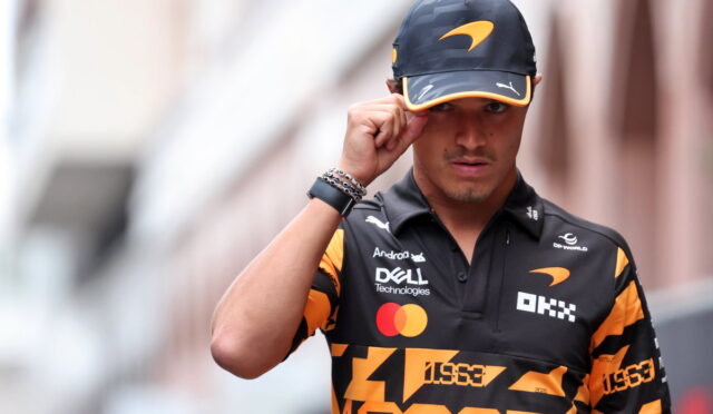 Male race car driver wearing a black and orange cap and team polo, adjusting his hat while walking toward the camera.