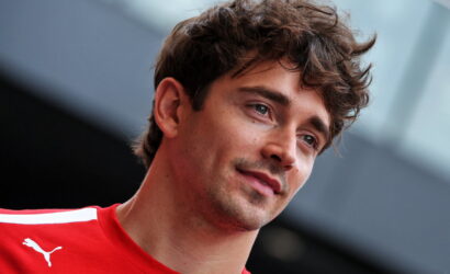 Portrait of a young man with tousled brown hair wearing a red sports jersey, looking slightly to the side, with a blurred background.
