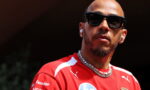 Close-up portrait of a man with black sunglasses, a red Ferrari racing shirt, and a pearl necklace wearing wireless earbuds in daylight.