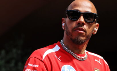 Close-up portrait of a man with black sunglasses, a red Ferrari racing shirt, and a pearl necklace wearing wireless earbuds in daylight.