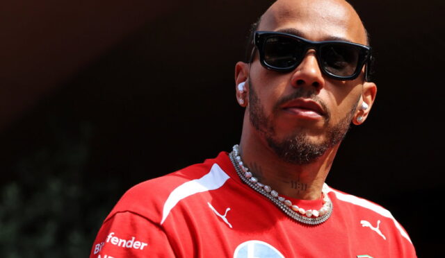 Close-up portrait of a man with black sunglasses, a red Ferrari racing shirt, and a pearl necklace wearing wireless earbuds in daylight.
