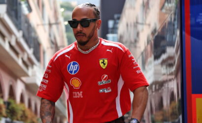 Man wearing a red Ferrari polo with sponsor logos, sunglasses, and chain walks down a city street.