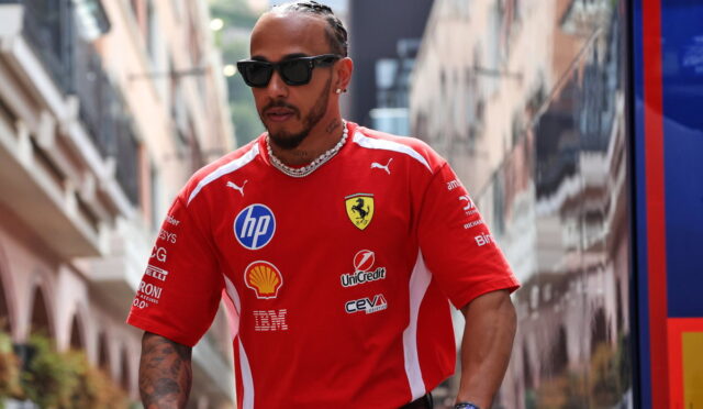 Man wearing a red Ferrari polo with sponsor logos, sunglasses, and chain walks down a city street.