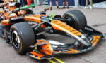 Orange Formula 1 race car in a pit/garage area with a driver in the cockpit and team personnel nearby, sponsors visible on the car.
