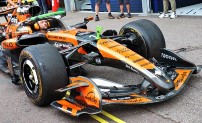 Orange Formula 1 race car in a pit/garage area with a driver in the cockpit and team personnel nearby, sponsors visible on the car.