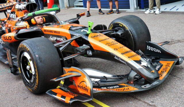 Orange Formula 1 race car in a pit/garage area with a driver in the cockpit and team personnel nearby, sponsors visible on the car.
