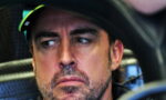 Close-up of a man with stubble wearing a racing helmet, looking to the left with a serious expression inside a car cockpit.