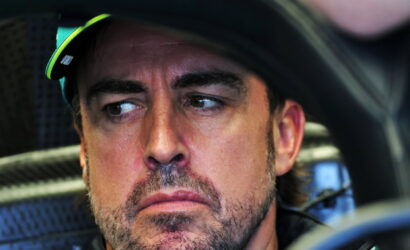 Close-up of a man with stubble wearing a racing helmet, looking to the left with a serious expression inside a car cockpit.