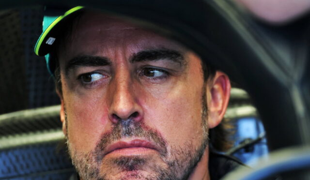 Close-up of a man with stubble wearing a racing helmet, looking to the left with a serious expression inside a car cockpit.