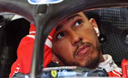 Close-up of a race car driver in a red suit inside a cockpit, looking upward with one hand resting on the head.