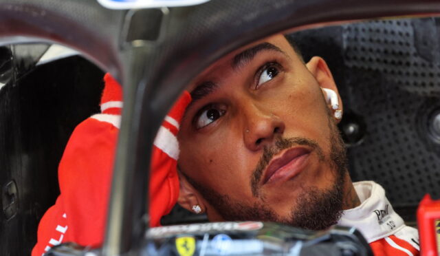 Close-up of a race car driver in a red suit inside a cockpit, looking upward with one hand resting on the head.