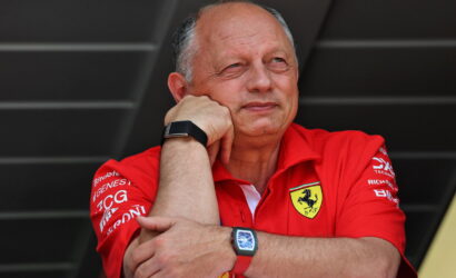 Man in a red Ferrari polo rests his chin on his folded arms, looking thoughtful against a dark background.”,