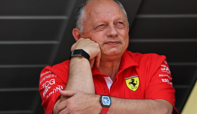 Man in a red Ferrari polo rests his chin on his folded arms, looking thoughtful against a dark background.”,
