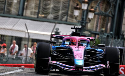 Front view of a pink-and-blue Formula 1 car speeding toward the camera on a race track, with spectators behind a fence in the background.