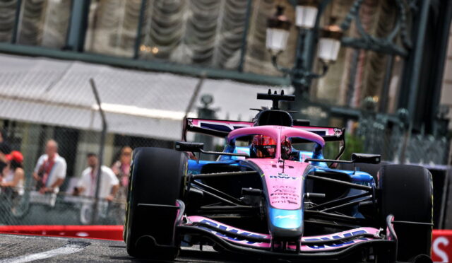 Front view of a pink-and-blue Formula 1 car speeding toward the camera on a race track, with spectators behind a fence in the background.