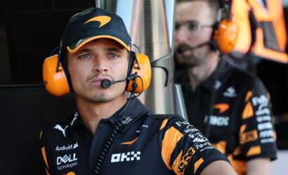 Two race team crew members in black and orange uniforms wearing large orange headsets in a pit area; the foreground member looks toward the camera while a teammate is blurred in the background.