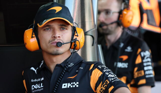 Two race team crew members in black and orange uniforms wearing large orange headsets in a pit area; the foreground member looks toward the camera while a teammate is blurred in the background.