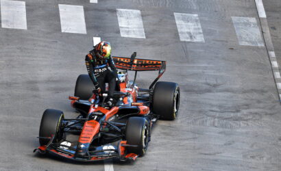 Formula 1 race car in orange and black livery taking a turn on a paved track, with the driver standing on the cockpit area.