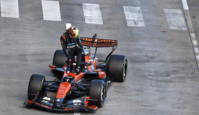 Formula 1 race car in orange and black livery taking a turn on a paved track, with the driver standing on the cockpit area.