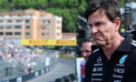 Man wearing a black polo with sponsor logos (Mercedes, Petronas, INEOS) grimaces as he stands at a race track, crowd in the background.