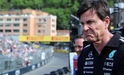 Man wearing a black polo with sponsor logos (Mercedes, Petronas, INEOS) grimaces as he stands at a race track, crowd in the background.