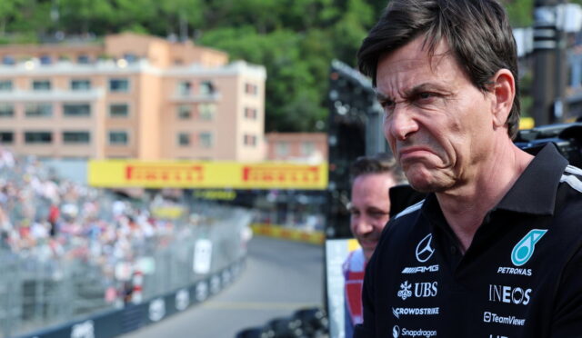 Man wearing a black polo with sponsor logos (Mercedes, Petronas, INEOS) grimaces as he stands at a race track, crowd in the background.