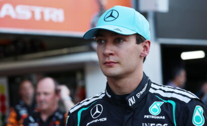 Male Formula 1 driver wearing a turquoise Mercedes cap and Petronas racing suit in the pit area.