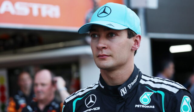 Male Formula 1 driver wearing a turquoise Mercedes cap and Petronas racing suit in the pit area.