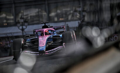 Pink and blue Formula 1 car on a city street circuit, driver in a red helmet, tires in motion.