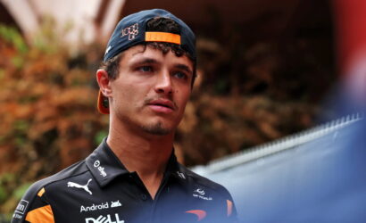 Young man in a backward orange cap and black sponsor polo, looking to the side outdoors at a race setting.