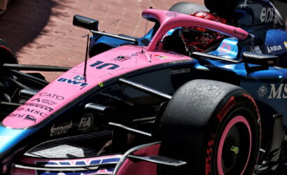 Pink and blue Formula 1 race car with sponsor logos on a brick-paved track area visible nearby surface.