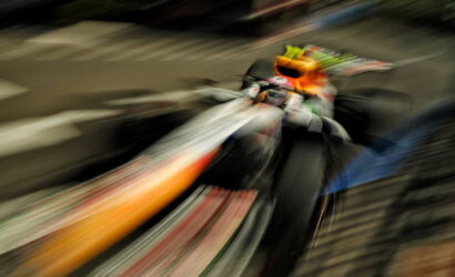 Motion-blurred photo of a motorcyclist with an orange helmet leaning into a turn on a racetrack, creating streaks of color behind the bike.