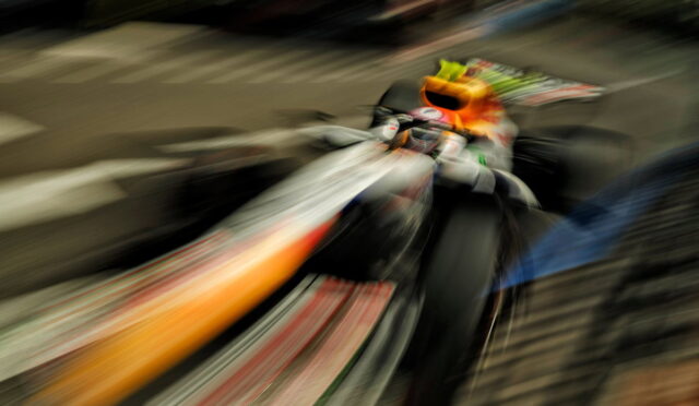 Motion-blurred photo of a motorcyclist with an orange helmet leaning into a turn on a racetrack, creating streaks of color behind the bike.