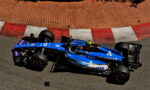 Blue Formula 1 car racing along a track with red and white curb, sponsor logos visible on the blue chassis (Atlassian).] ,