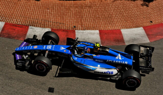 Blue Formula 1 car racing along a track with red and white curb, sponsor logos visible on the blue chassis (Atlassian).] ,