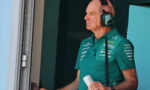 Pit crew member in a teal polo and headset stands at a pit window, holding a white can.