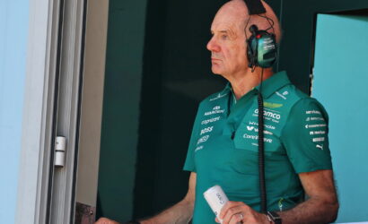 Pit crew member in a teal polo and headset stands at a pit window, holding a white can.