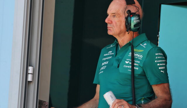 Pit crew member in a teal polo and headset stands at a pit window, holding a white can.