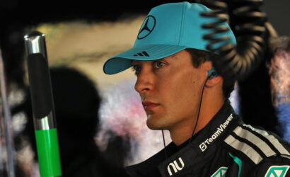 Side profile of a race driver wearing a teal cap and TeamViewer racing suit, listening with an in-ear headset in a pit area