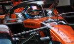 Close-up of an orange Formula 1 car cockpit with a driver wearing a blue helmet, sponsor logos like Richard Mille visible on the chassis, and race equipment surrounding the car.