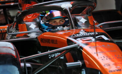 Close-up of an orange Formula 1 car cockpit with a driver wearing a blue helmet, sponsor logos like Richard Mille visible on the chassis, and race equipment surrounding the car.