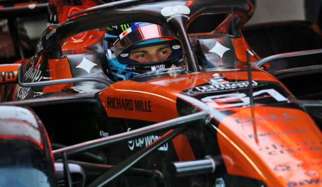Close-up of an orange Formula 1 car cockpit with a driver wearing a blue helmet, sponsor logos like Richard Mille visible on the chassis, and race equipment surrounding the car.