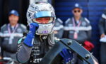 Formula 1 driver in a Petronas helmet and blue gloves holds a car component while team members watch in the background.
